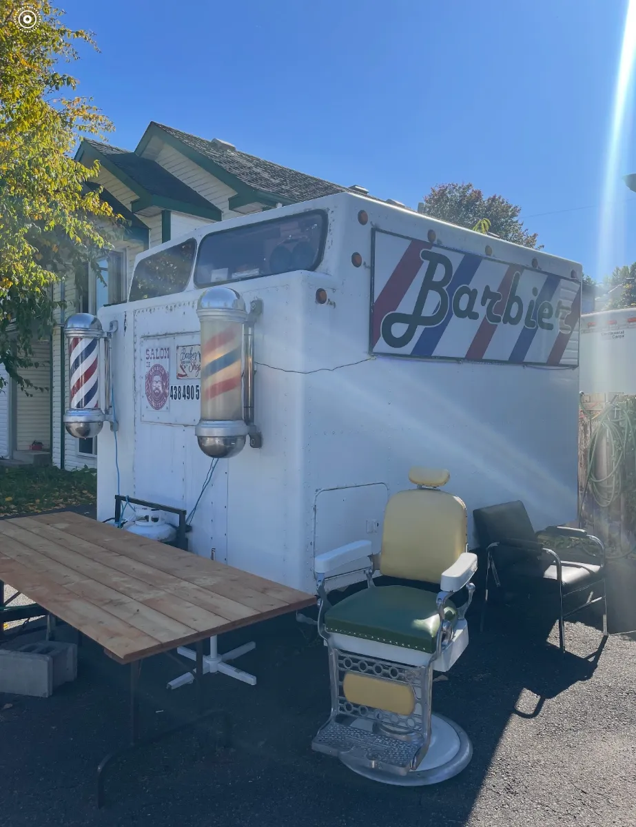 Le salon mobile avec son enseigne Barbier, ses poteaux et un fauteuil de barbier d'époque devant