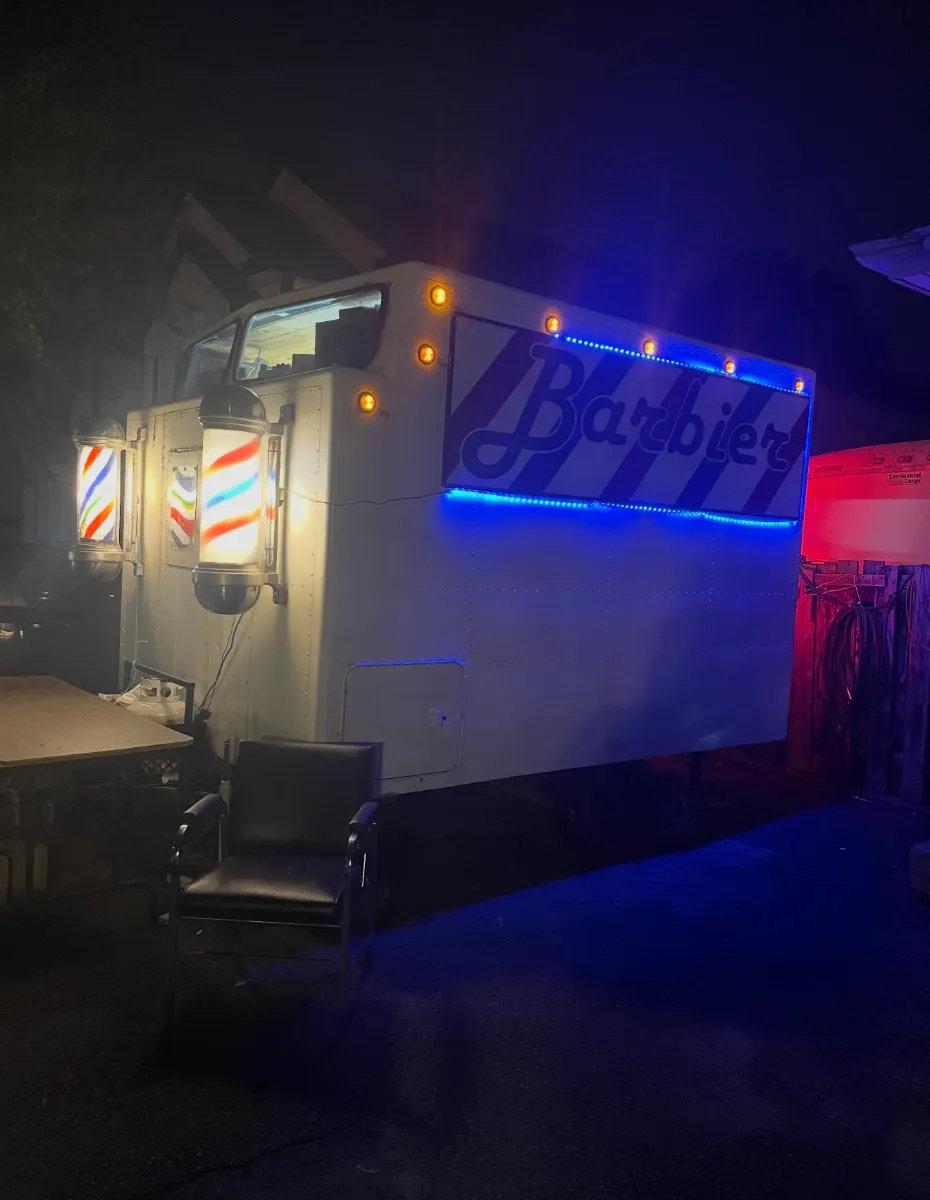 Le salon mobile le soir, poteaux de barbier allumés et enseigne Barbier éclairée en bleu